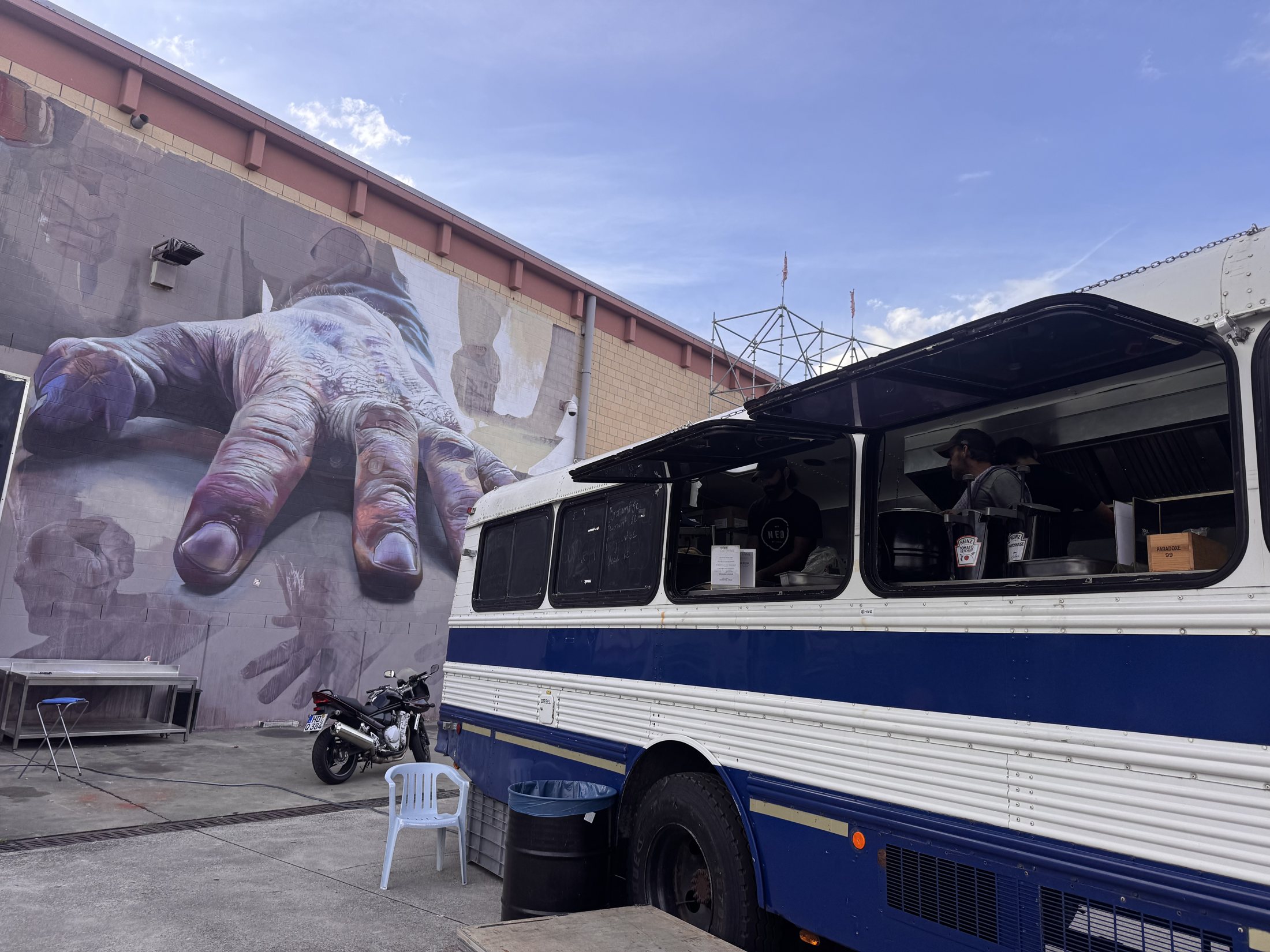 Der weiß-blaue BAM! Bus mit offenen Klappen vor einem großen Wandbild, die Crew kocht im Fenster